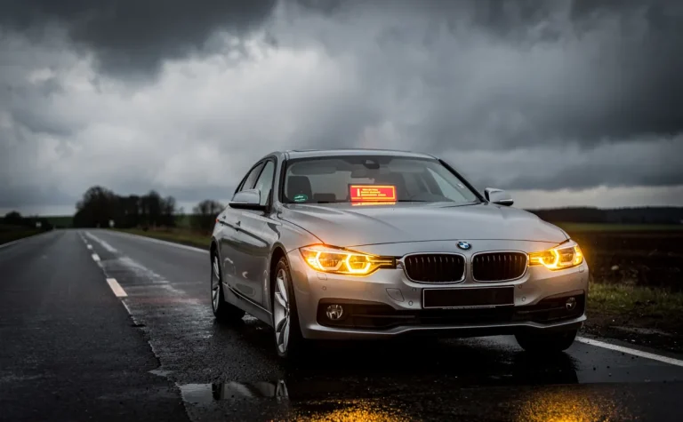 Une BMW stationnée sur le bord de la route avec les feux de détresse allumés et un message d’alerte visible sur le tableau de bord, sous un ciel nuageux.