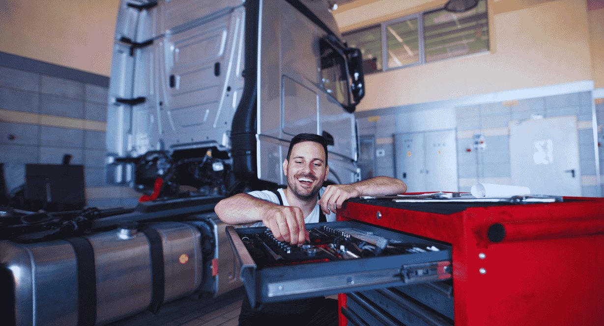 Mécanicien souriant qui choisit un outil dans une servante rouge, devant la cabine d'un camion en atelier